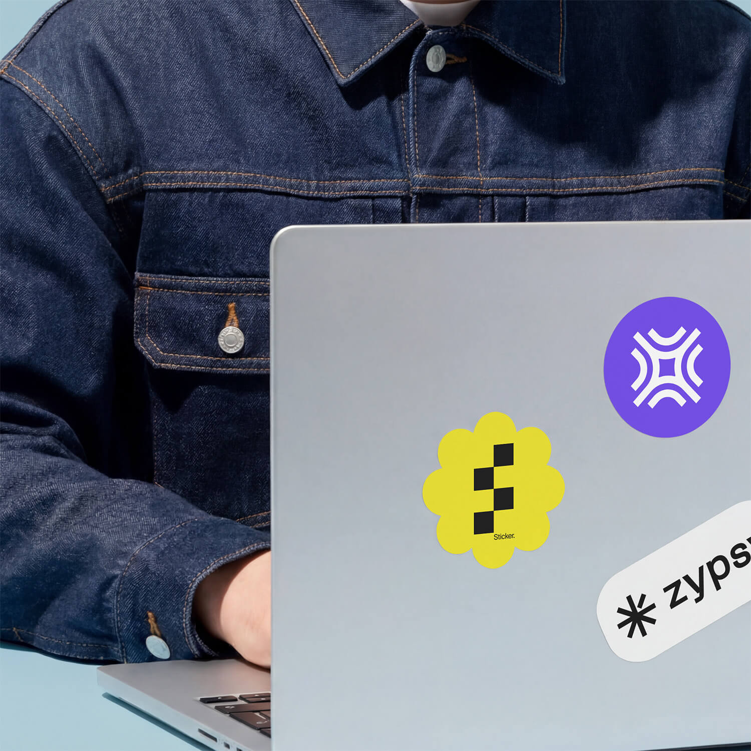 Person in denim jacket using a laptop with yellow and purple logo stickers and a white Zypsa sticker.