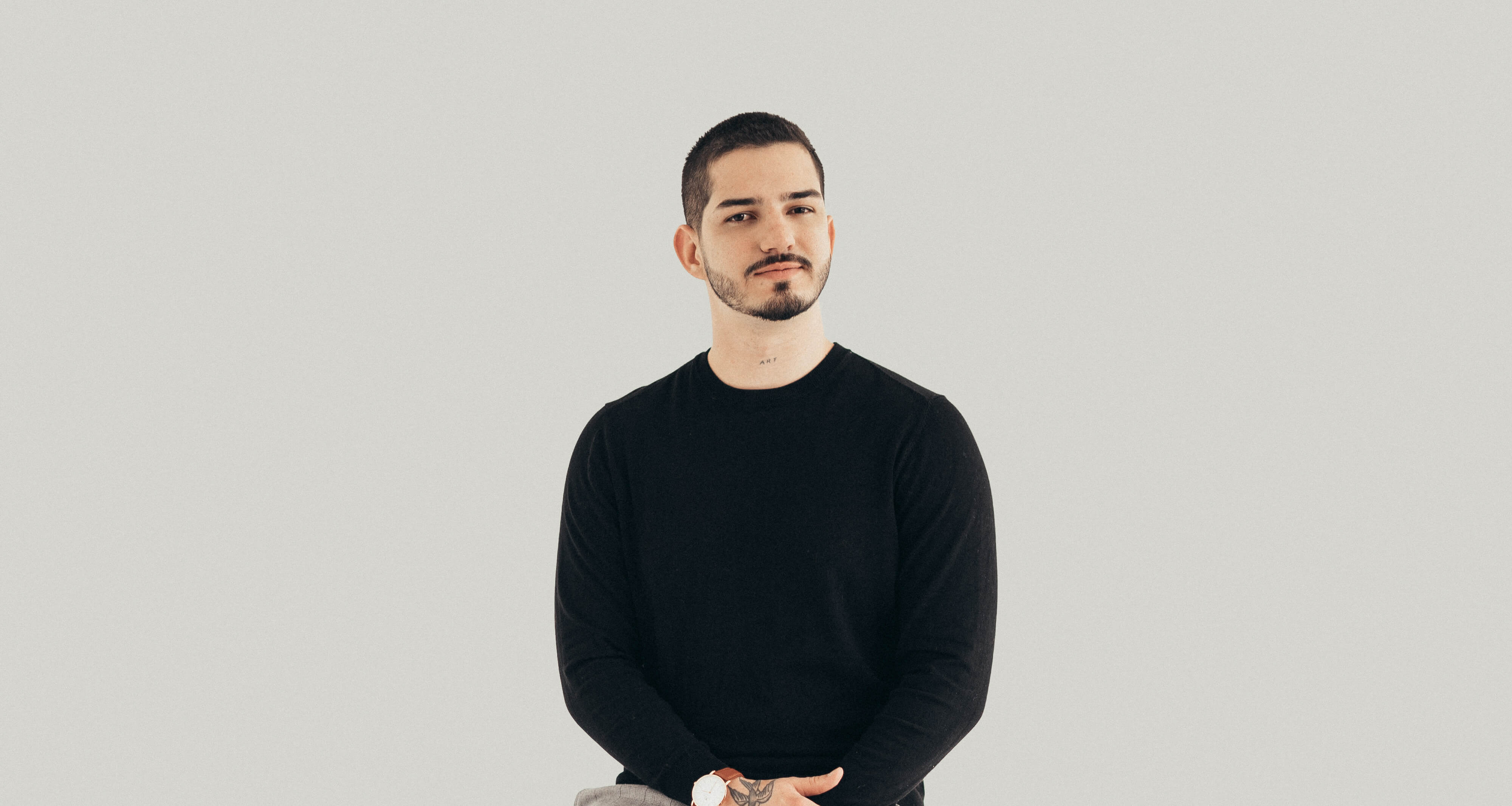Man with short hair and beard wearing black sweater sitting with hands folded on light background.
