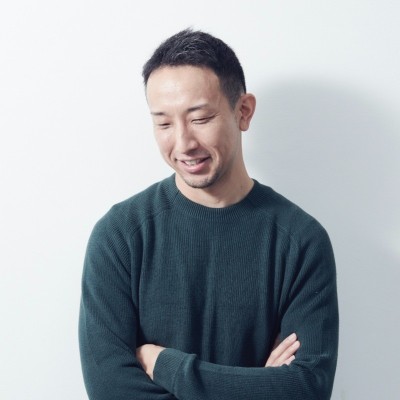 Man with short black hair wearing a dark green sweater, smiling with arms crossed, against a white wall.