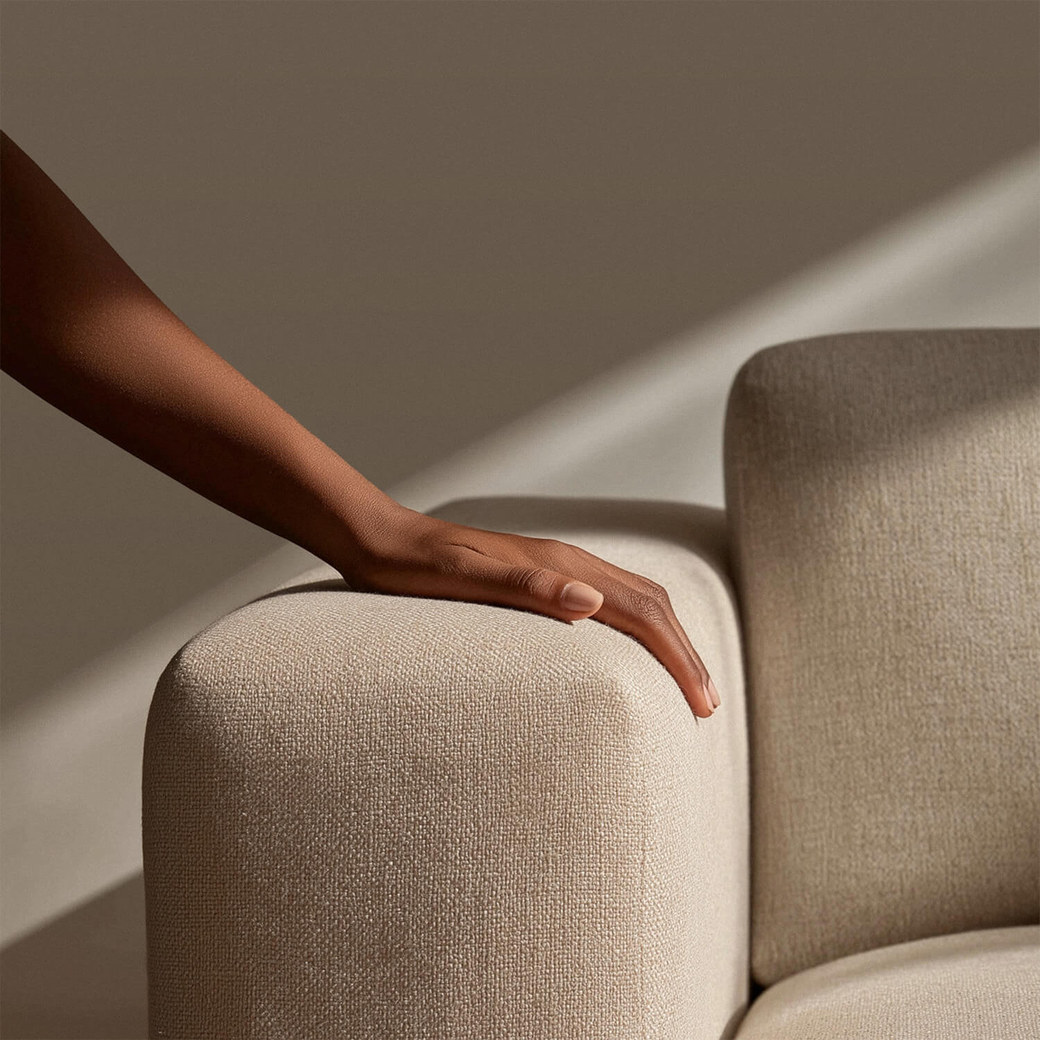 Hand resting on the arm of a beige textured sofa with sunlight casting shadows nearby.