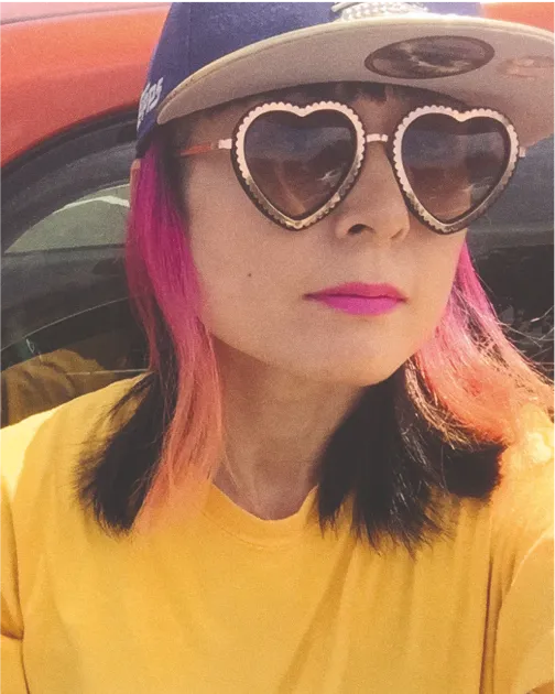 Person with pink hair wearing heart-shaped sunglasses, a cap, and a yellow shirt in a car.